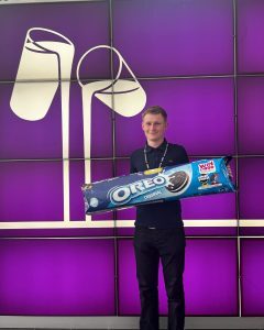 Joe, a light-skinned male student, stands smiling in front of a large purple Mondelēz display wall, holding an oversized pack of Oreo biscuits.