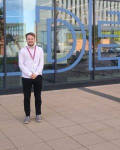 Michael, a light-skinned male student, stands outside the Office for National Statistics building wearing a white shirt and lanyard.