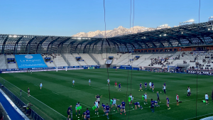 Players warming up on a rugby pitch inside a large stadium, with snow-covered mountains visible in the background.