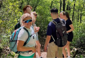 A group of people standing on a sunny forest trail with backpacks, pausing during a hike among dense green trees.