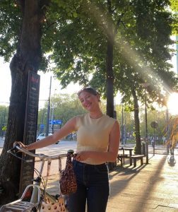Olivia holding a bicycle by the handlebars, standing on a tree-lined street on a sunny day in Rotterdam.