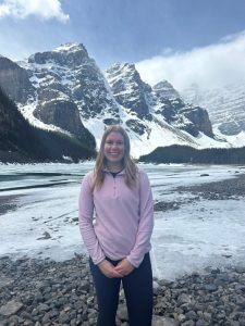 Tamsin standing on a rocky lakeshore with a frozen lake and tall snow-covered mountains in the background.