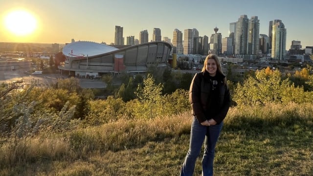 Tamsin standing on a grassy hill at sunset with a large arena and the Calgary skyline in the background.