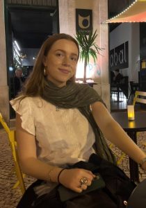 Wendy sitting at an outdoor café table in the evening, with warm lighting, yellow chairs and a drink on the table.