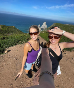 Wendy and a friend taking a selfie on a cliffside path overlooking the coastline, with blue sea and rock formations in the background.