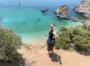 Wendy standing on a cliff above a sandy beach with clear turquoise water and rock formations below.