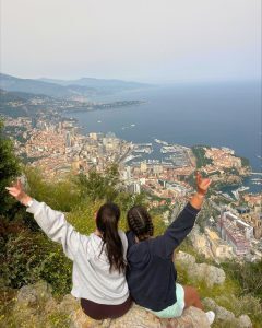 Two students sitting on a rock overlooking the coastline and cityscape of Nice, France, with their arms raised in celebration