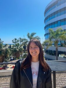 Abi standing outdoors on a sunny terrace with palm trees and a modern glass building behind her.