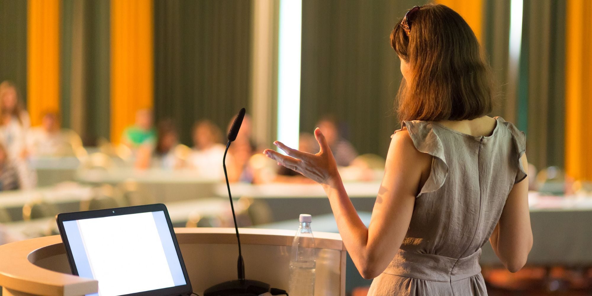 Woman giving a presentation at a lectern, gesturing towards an audience seated in a conference room.